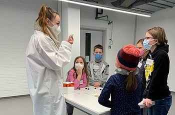 Eine Person mit Maske erklärt den Kindern ein Experiment