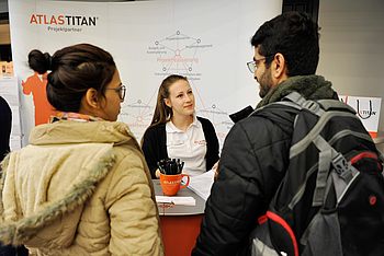 Zwei Personen fragen eine Person, die einen Stand auf einer Messe betreibt