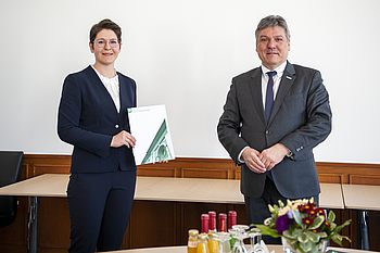 Eine Person hält ein Dokument in der Hand und steht neben einer Person 